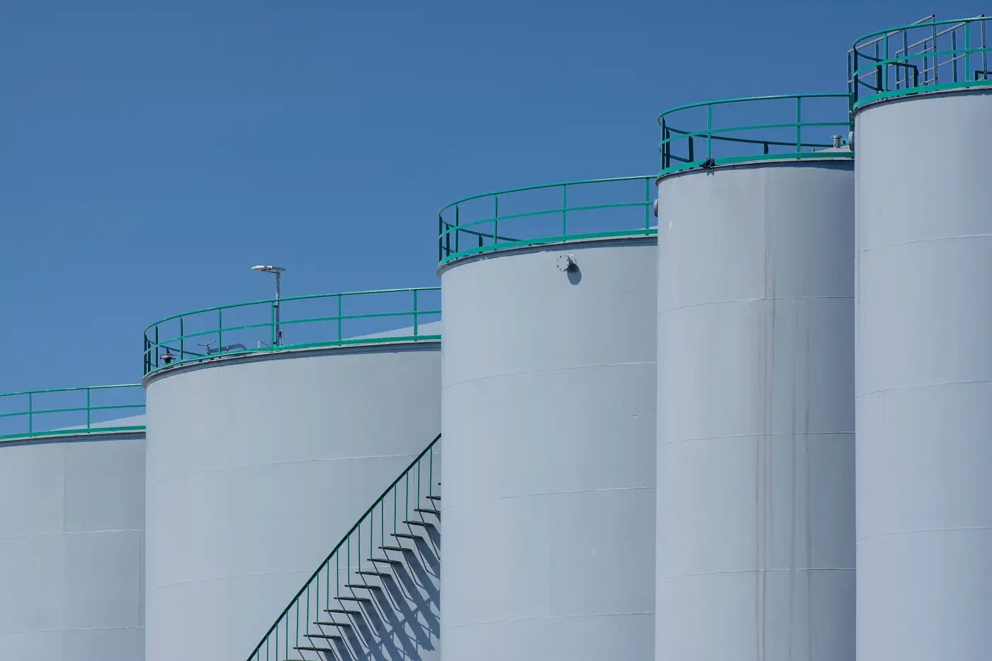 Close view of industrial storage tank tops with green perimeter railings and a stairway, set against a clear blue sky.